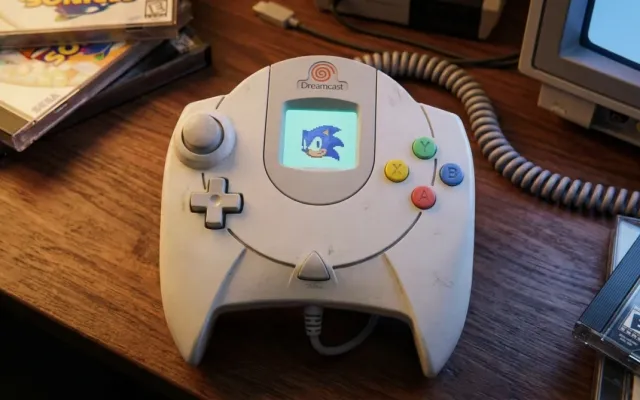 Controle branco clássico do Sega Dreamcast exibindo a tela LCD do VMU acoplada no centro do joystick.