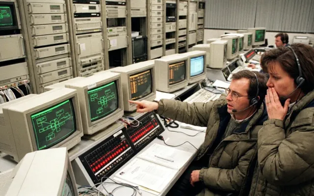 Sala de controle de telecomunicações dos anos 1990 com monitores exibindo alertas da rede AT&T.